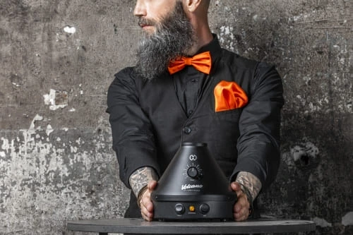 Ein Mann mit Mattschwarzen Hemd und Orangener Fliege präsentiert vor sich den Volcano Classic Vaporizer in der ONYX Edition